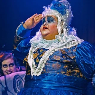 Performer in elaborate blue and gold costume with white wig saluting on stage